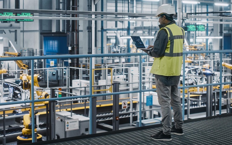 Male engineer in high visibility vest using laptop computer looking at automotive manufacturing facility